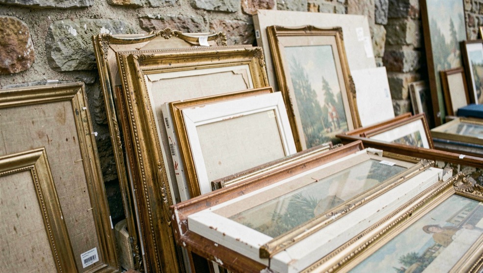 Étal de brocante avec des tableaux empilés contre un mur de pierre, cadres dorés patinés et toiles non encadrées mêlées