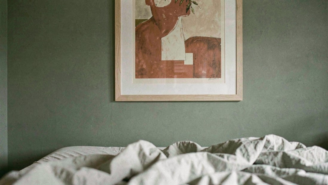 Chambre aux murs vert sauge avec un tableau aux tons terracotta et crème encadré de chêne clair au-dessus d’un drap froissé